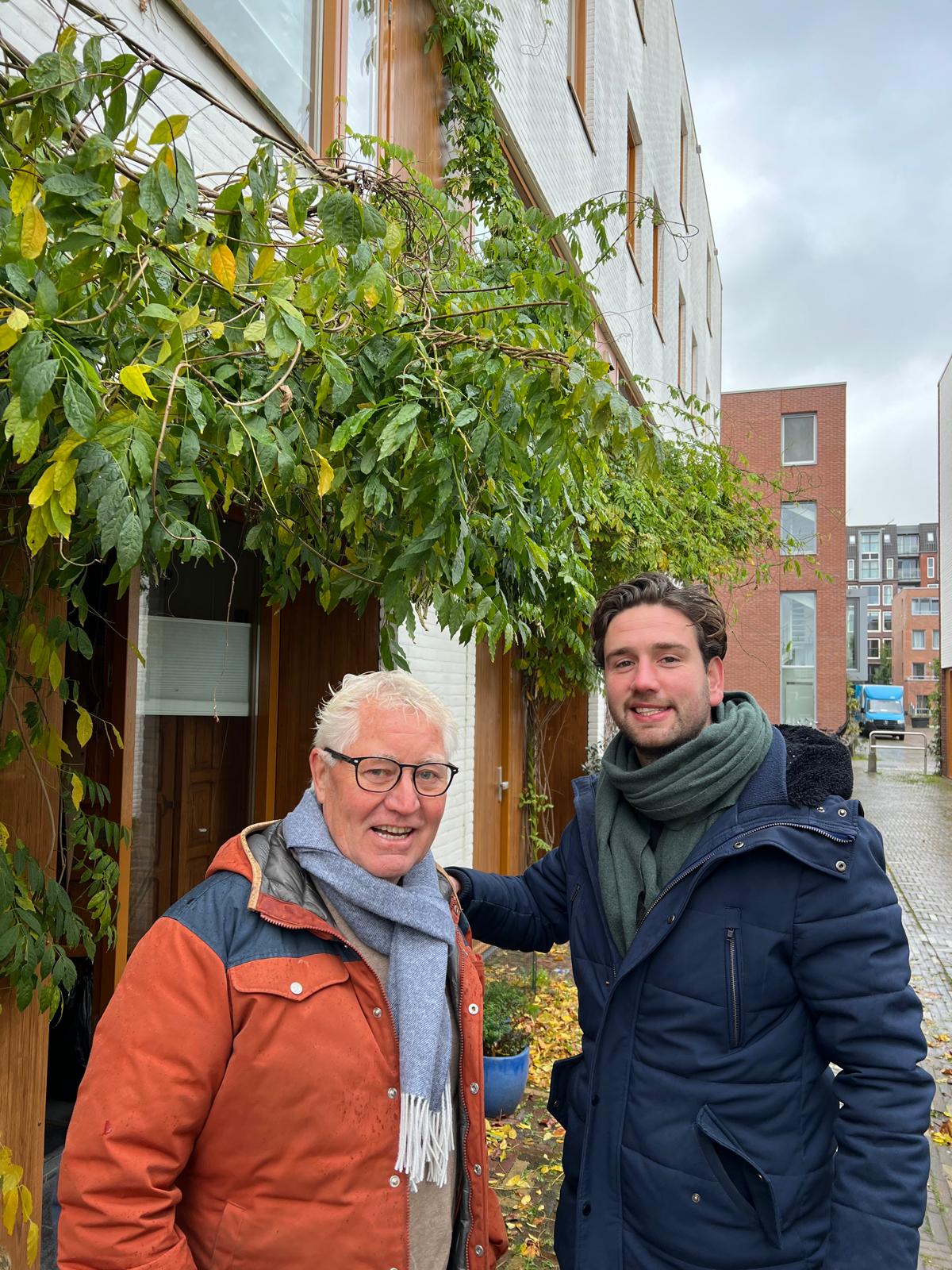 Een oudere man en een jonge man staan voor een gebouw met veel planten op de gevel