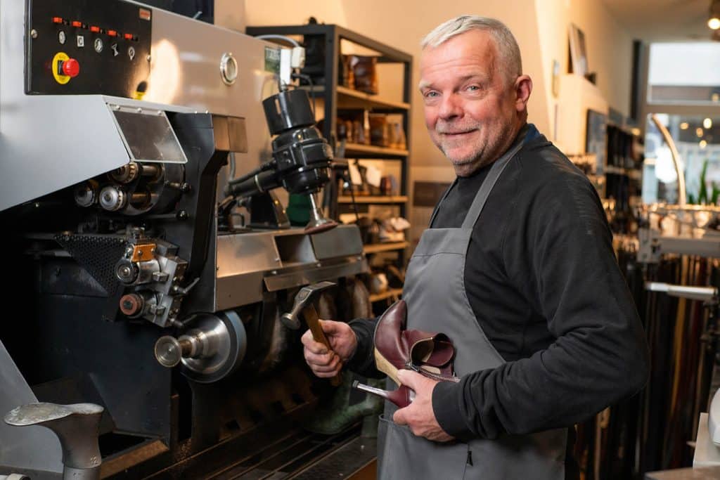 Schoenmaker met een schoen en hamer in zijn handen voor het apparaat waar hij de schoenen mee maakt.