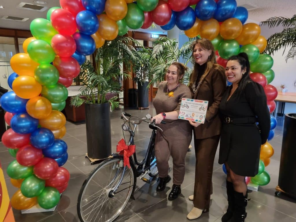 Burgemeester Anja Schouten met een vrouw die een fiets krijgt en een vrouw van project Fiets. Ze staan onder een ballonnenboog.